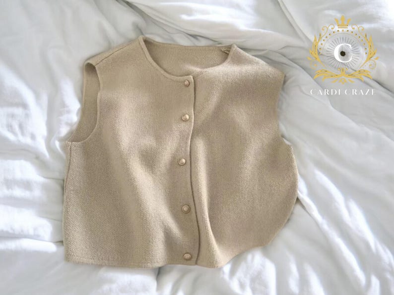May include: A beige, sleeveless sweater vest with a round neckline and a button-down front. The vest has a soft texture and is buttoned with six small, round buttons. The garment is displayed on a white, textured surface.