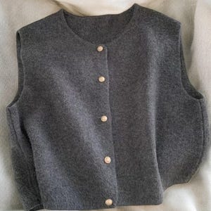 May include: A gray sleeveless vest with a round neckline and four gold-colored buttons down the front. The vest is made of a soft, textured fabric and appears to be a comfortable layering piece.