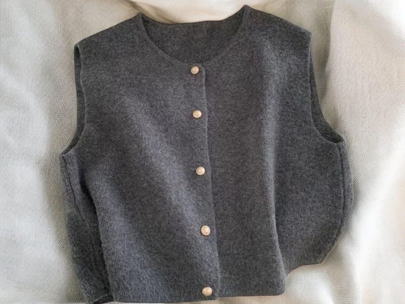 May include: A gray sleeveless vest with a round neckline and four gold-colored buttons down the front. The vest is made of a soft, textured fabric and appears to be a comfortable layering piece.