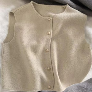 May include: A beige, sleeveless sweater vest with a round neckline and a button-down front. The vest is made of a soft, knitted material and features four round, pearl-like buttons. The garment is lying flat on a white and gray surface.