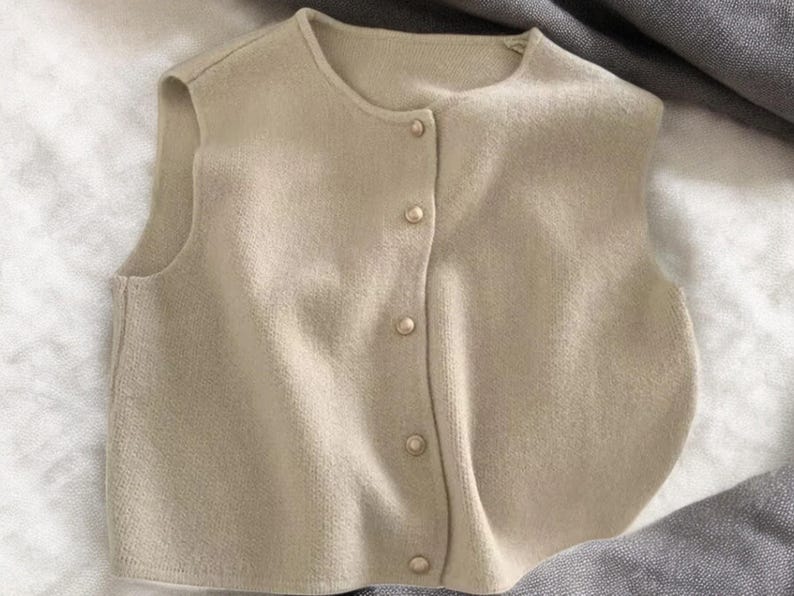 May include: A beige, sleeveless sweater vest with a round neckline and a button-down front. The vest is made of a soft, knitted material and features four round, pearl-like buttons. The garment is lying flat on a white and gray surface.