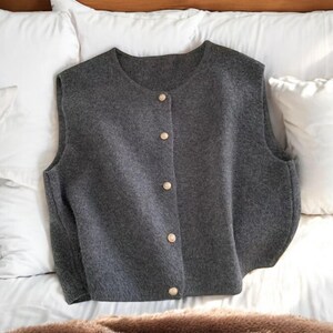 May include: A gray, button-down vest with a round neckline. The vest has a soft, textured appearance and is fastened with five gold-colored buttons. The garment is displayed on a white surface, suggesting a focus on apparel.