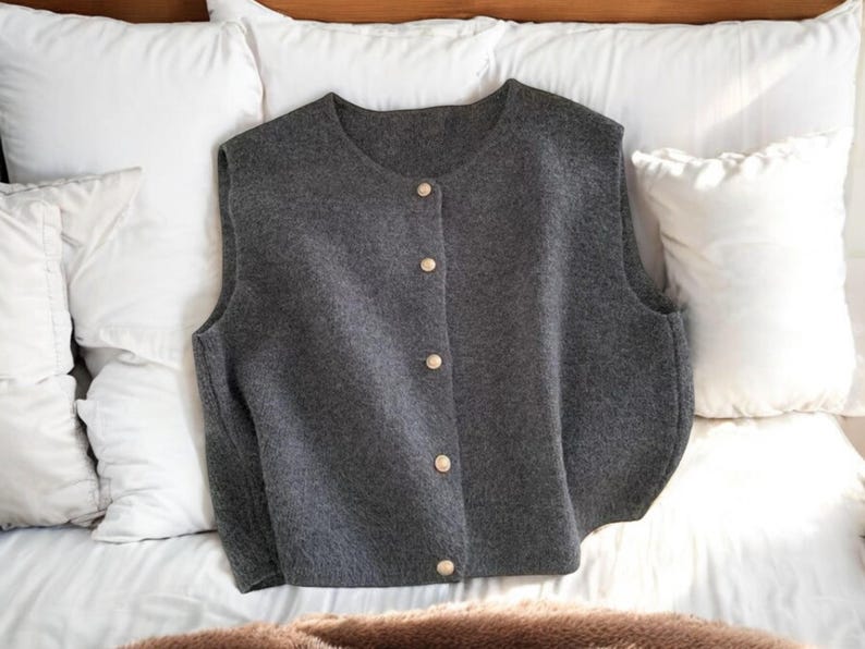 May include: A gray, button-down vest with a round neckline. The vest has a soft, textured appearance and is fastened with five gold-colored buttons. The garment is displayed on a white surface, suggesting a focus on apparel.