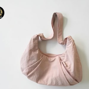 Pode incluir: Uma bolsa de ombro rosa claro com uma única alça. A bolsa tem uma forma curva com pregas e um fecho de zíper. A bolsa é feita de um material de tecido macio. O logotipo "CARDI CRAZE" está em um círculo preto com uma coroa dourada.