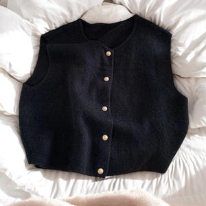 May include: A navy blue vest with a round neckline and a button-down front. The vest has a textured appearance and is adorned with five gold-colored buttons. The garment is displayed on a white, textured surface.