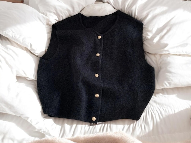 May include: A navy blue vest with a round neckline and a button-down front. The vest has a textured appearance and is adorned with five gold-colored buttons. The garment is displayed on a white, textured surface.