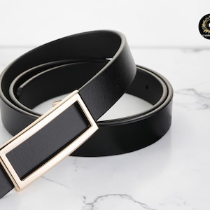 May include: A black belt with a rectangular gold-colored buckle. The belt is made of a smooth, dark material and is coiled on a white marble surface. The buckle has a black center.