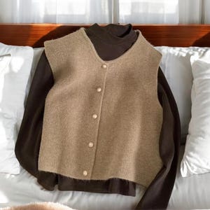 May include: A tan vest with gold buttons layered over a dark brown long-sleeved shirt. The vest has a V-neck and a slightly textured appearance. The clothing is displayed on a bed with white pillows.