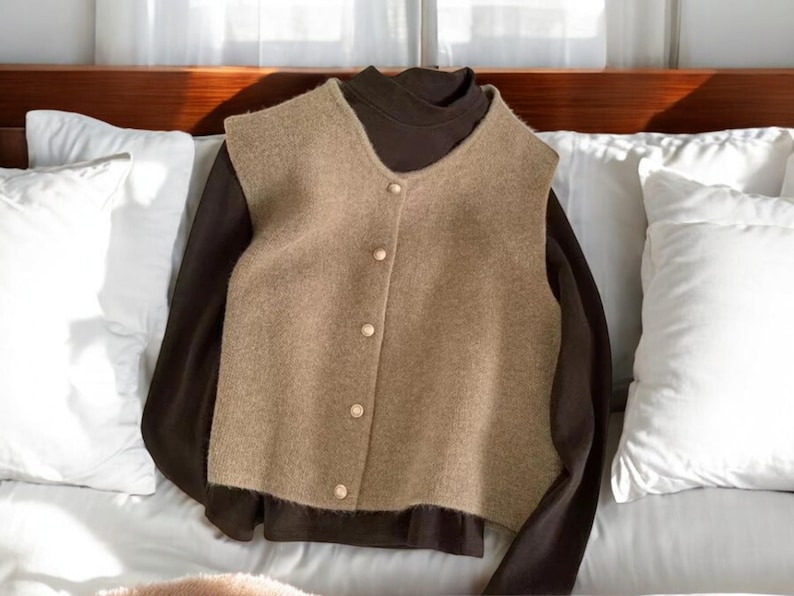 May include: A tan vest with gold buttons layered over a dark brown long-sleeved shirt. The vest has a V-neck and a slightly textured appearance. The clothing is displayed on a bed with white pillows.