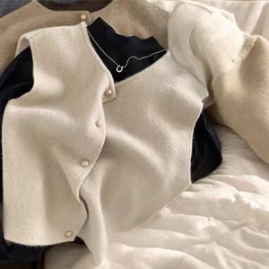 May include: A collection of neutral-toned sweaters and vests. A cream-colored vest with gold buttons is layered over a black turtleneck and a silver necklace. Other sweaters are in shades of gray, brown, and beige. The items are arranged on a white, textured surface.