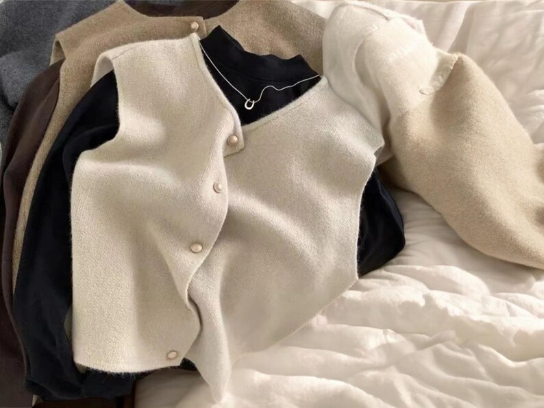 May include: A collection of neutral-toned sweaters and vests. A cream-colored vest with gold buttons is layered over a black turtleneck and a silver necklace. Other sweaters are in shades of gray, brown, and beige. The items are arranged on a white, textured surface.