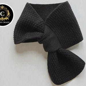 May include: A black scarf with a textured, waffle-knit design. The scarf is knotted in the center, creating a stylish and warm accessory. The Cardi Craze logo is in the upper left corner.