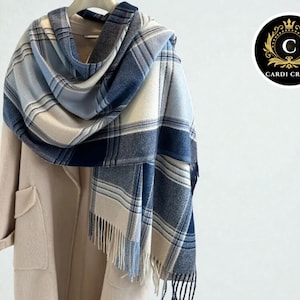 May include: A blue and cream plaid scarf with fringe is draped over a beige coat. The scarf features a mix of light and dark blue squares and rectangles. The coat has large pockets and a simple design. The scarf is hanging on a hanger.