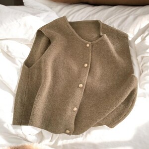 May include: A sleeveless, button-down vest in a muted olive green hue. The vest features a round neckline and is fastened with five gold-colored buttons. The garment is made of a soft, textured material and is displayed on a white bedspread.