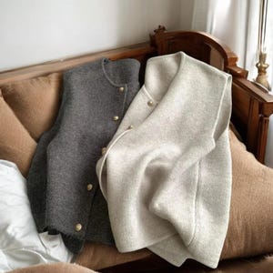 May include: Two wool vests, one gray and one off-white, with gold-colored buttons, rest on a brown cushioned surface. The vests are buttoned and appear to be made of a soft, textured material. The background includes a wooden headboard and a window.