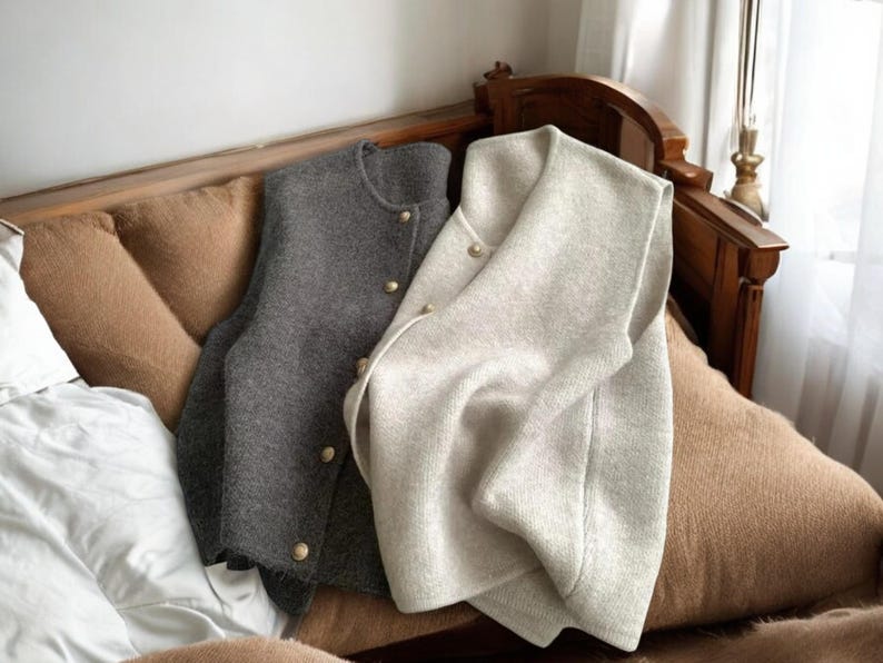 May include: Two wool vests, one gray and one off-white, with gold-colored buttons, rest on a brown cushioned surface. The vests are buttoned and appear to be made of a soft, textured material. The background includes a wooden headboard and a window.