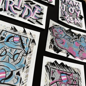 Könnte beinhalten: Eine Sammlung von Aufklebern mit Text und Illustrationen. Das Wort „PRIDE“ ist in großen Buchstaben mit blauer, rosa und weißer Farbe gehalten. Andere Aufkleber sagen „TRANS JOY IS REAL“ und „YOU ARE LOVED“. Spielkarten-Designs sind ebenfalls enthalten.