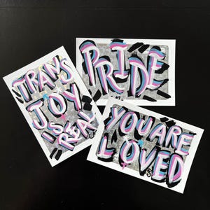 Puede incluir: Tres pegatinas rectangulares con texto manuscrito sobre un fondo holográfico plateado. Las pegatinas presentan las frases "TRANS JOY IS REAL", "PRIDE" y "YOU ARE LOVED" en rosa, azul y negro, en un estilo graffiti.
