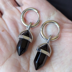 Op de afbeelding: Een paar zilveren oorringen met zwarte, puntige kristallen hangers. De kristallen zijn gezet in zilver metaal. De oorbellen worden getoond tegen een neutrale achtergrond.