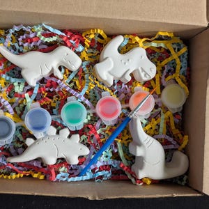 Könnte beinhalten: Ein Dino-Malset in einem Karton. Das Set enthält vier weiße Dinosaurierfiguren, sechs kleine Farbtöpfe in verschiedenen Farben und einen Pinsel. Die Box ist mit bunten Papierschnipseln gefüllt.