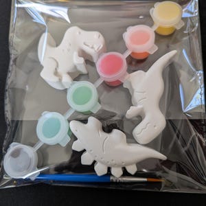Pode incluir: Um kit de pintura com tema de dinossauros é apresentado em um saco plástico transparente. O kit inclui quatro figuras de dinossauros brancos, seis potes de tinta pequenos de várias cores e um pincel azul. Os dinossauros são um T-Rex, um Estegossauro e duas outras espécies não identificadas.