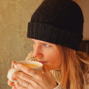 Op de afbeelding: Een persoon met een zwarte gebreide beanie en een crèmekleurige kabeltrui houdt een witte koffiemok vast. De persoon draagt twee zilveren ringen. De achtergrond is een getextureerde muur.