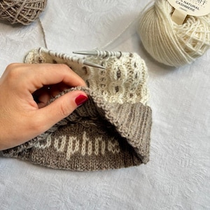 Op de afbeelding: Een gedeeltelijk voltooide gebreide muts in crème- en bruintinten, met de naam "Daphne" zichtbaar op de omgevouwen rand. Twee bollen wol en breinaalden zijn ook aanwezig. Het garen is gelabeld "Cardiff Classic 100% Natural Cashmere."