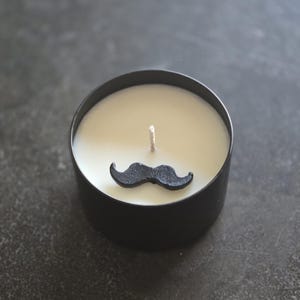 May include: A black candle in a round black container with a black mustache accent. The candle has a white wax and a single wick. The candle is on a dark gray surface.