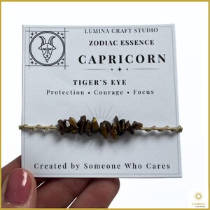 May include: A bracelet featuring brown tiger's eye stone chips on a beige cord, attached to a white display card labeled Lumina Craft Studio, Zodiac Essence, Capricorn, Tiger's Eye, Protection, Courage, Focus, Created by Someone Who Cares.