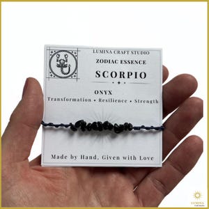 May include: A dark blue string bracelet featuring small, irregular black onyx stones. The bracelet is attached to a white display card labeled Lumina Craft Studio Zodiac Essence Scorpio, listing the words Transformation, Resilience, and Strength.