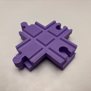 May include: A solid purple, cross-shaped puzzle piece with rounded edges and interlocking features. The piece has a textured surface and is designed to connect with other similar pieces. The object is made of a solid material.