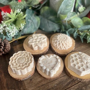Pode incluir: Seis pequenos sabonetes decorativos brancos com diferentes desenhos, incluindo uma flor, um coração, um favo de mel e um padrão de redemoinho. Os sabonetes são exibidos em pequenas rodadas de madeira.