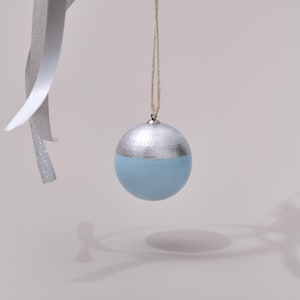 Op de afbeelding: Een lichtblauwe en zilveren kerstbal met een touwtje om op te hangen.