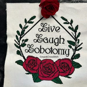 May include: White cross-stitch embroidery with a black diamond frame and the text "Live Laugh Lobotomy" in a cursive font. The diamond frame is surrounded by green leaves and three red roses.