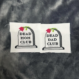 Op de afbeelding: Twee kruissteek-borduurwerken die grafstenen voorstellen met de tekst "Dead Mom Club" en "Dead Dad Club". Elke grafsteen heeft een rode roos erop.