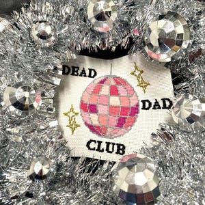 May include: A white cross-stitch design with a pink disco ball, gold stars, and the text "Dead Dad Club". The design is surrounded by silver tinsel.