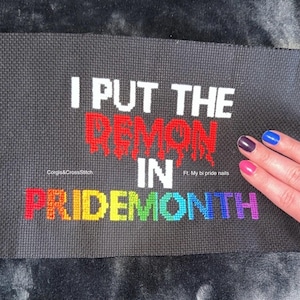 Puede incluir: Tela negra bordada a punto de cruz con el texto "I PUT THE DEMON IN PRIDEMONTH". La palabra "DEMON" está en rojo, y "PRIDEMONTH" está en colores arcoíris. El texto "Corgis&CrossStitch" también es visible.