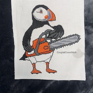 May include: A cross-stitch embroidery on white fabric featuring a puffin with black, white, and orange feathers holding an orange chainsaw. The text Corgis&CrossStitch is embroidered in black on the right side of the puffin's body.