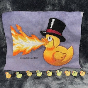 Op de afbeelding: Kruissteekkunst met een gele eend met een zwarte hoge hoed met een rode band, die vlammen uitblaast. De tekst "Corgis&CrossStitch" is zichtbaar. Kleine gele eendenfiguurtjes zijn eronder geplaatst.