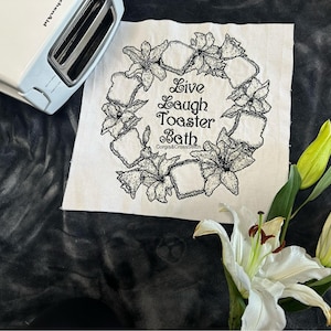 Puede incluir: Una tostadora KitchenAid blanca junto a un cuadrado de tela blanca con un diseño de corona floral y las palabras "Live Laugh Toaster Bath". Lirios blancos con puntas amarillas en primer plano.