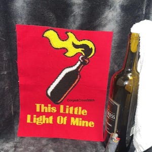 Op de afbeelding: Een rood kruissteekstuk met een flesillustratie en de tekst "This Little Light Of Mine". Een wijnfles, een aansteker en een witte handdoek liggen naast de kruissteek.