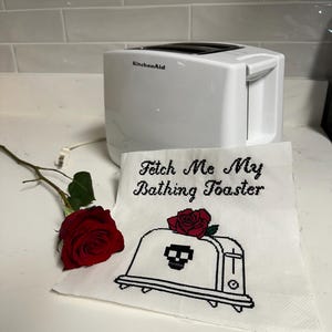 Op de afbeelding: Een witte KitchenAid-toaster met een witte geborduurde keukendoek met de tekst "Fetch Me My Bathing Toaster" en een geborduurd zwart-wit beeld van een toaster met een schedel en een rode roos erop.