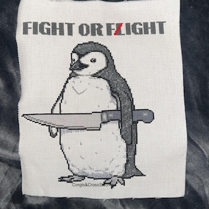 May include: Cross-stitch embroidery of a penguin holding a knife with the text "Fight or Flight" above it. The embroidery is on white fabric with black and gray thread.