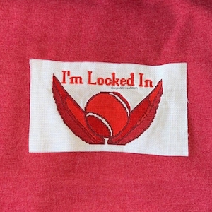 May include: A white cross-stitch patch with the text "I'm Locked In" in red, along with a red tennis ball design and wings. The patch is set against a red fabric background. The text "Corgis & CrossStitch" is also visible.