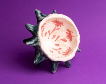 Puede incluir: Un cuenco de cerámica con un exterior verde y un interior blanco con diseños florales rojos. El cuenco tiene una forma puntiaguda en forma de estrella.