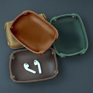 May include: Four leather catchall trays in shades of brown and green are displayed on a dark surface. One tray contains white earbuds. The trays are rectangular with rounded corners, stitched edges, and brass-colored rivets.