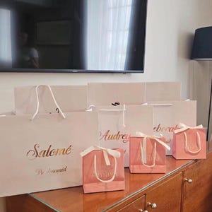 May include: Several white and pink gift bags with gold lettering and white ribbon handles are arranged on a wooden surface. The bags are labeled with names like "Salom&eacute;" and "Audrey". The smaller pink bags have the name "Amaya" printed on them.