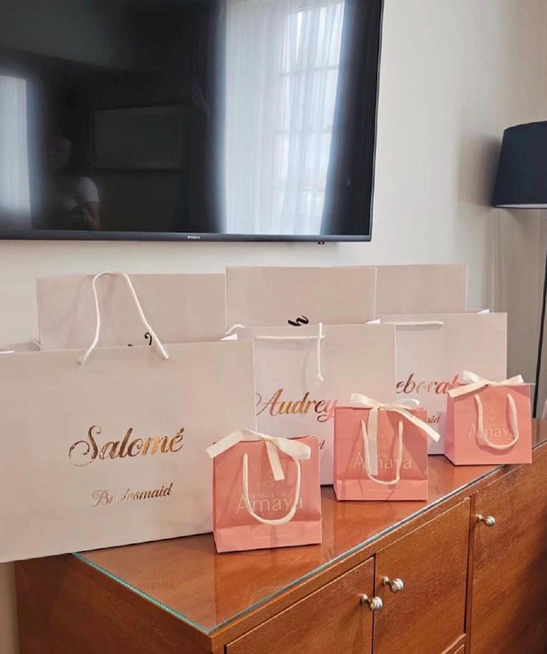May include: Several white and pink gift bags with gold lettering and white ribbon handles are arranged on a wooden surface. The bags are labeled with names like "Salom&eacute;" and "Audrey". The smaller pink bags have the name "Amaya" printed on them.