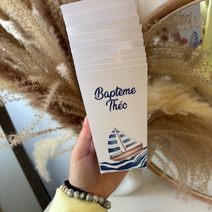 Peut inclure: Une pile de boîtes en papier blanc avec un motif de voilier bleu et blanc. La boîte porte l'inscription "Bapteme Theo".