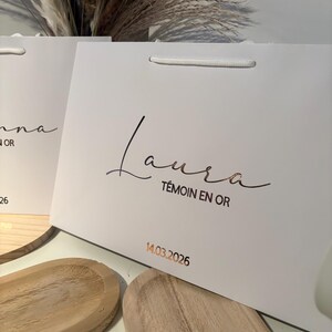 May include: White gift bag with a gold-colored script that reads "Laura" and "T&eacute;moin en Or". The date "14.03.2026" is printed in gold. The bag has a white cord handle.